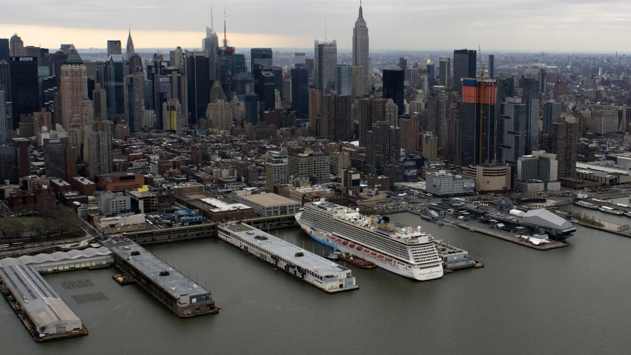 Aerial view of a cruise ship docked at the Manhattan Cruise Terminal piers on the Hudson River, with the Midtown Manhattan skyline and the Empire State Building behind